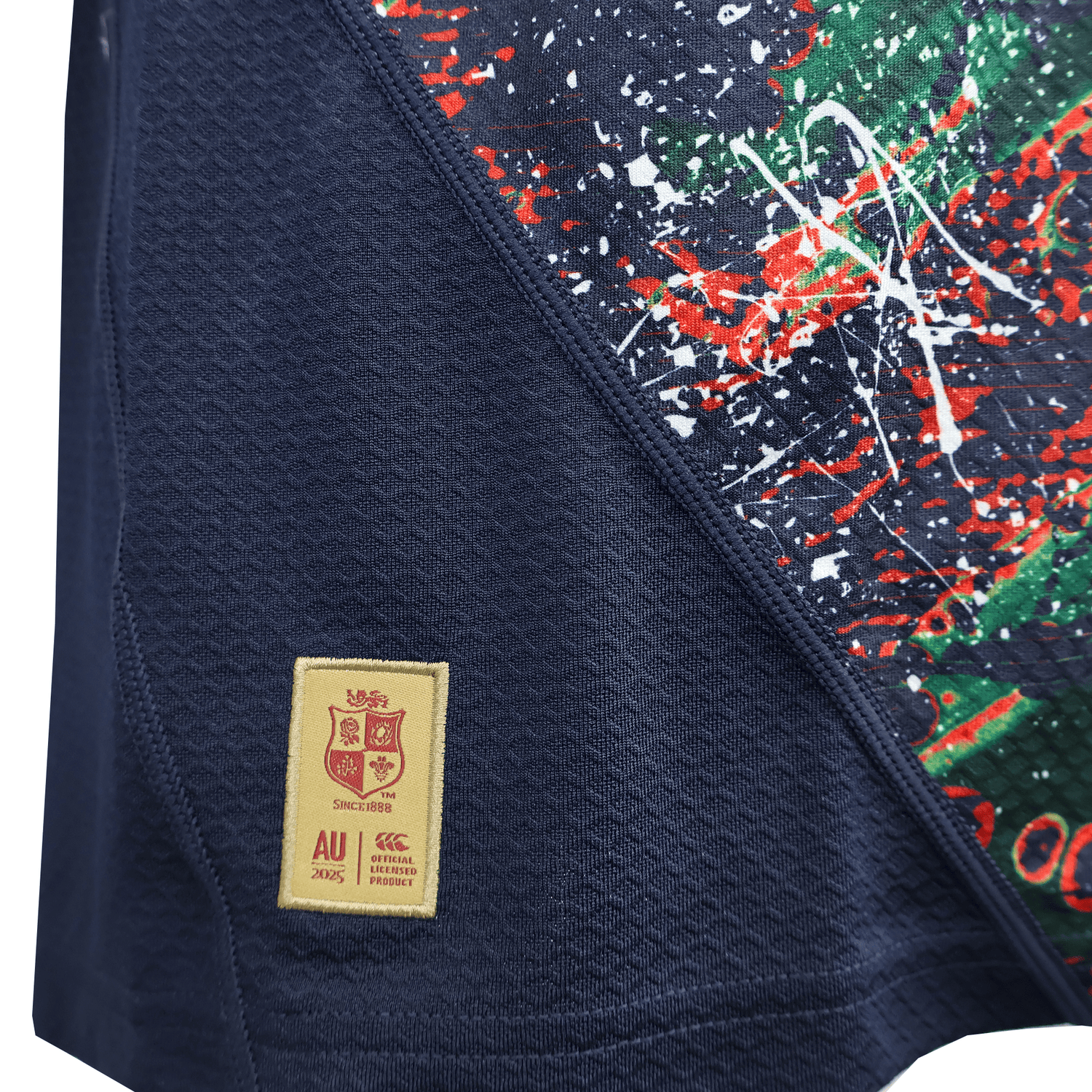 A close-up of the British & Irish Lions 2025 Replica Warm Up Jersey by Canterbury in blue athletic fit, featuring a vibrant splatter design on one side and a stitched patch with crest and text on the other. The moisture-wicking fabric ensures comfort throughout wear.