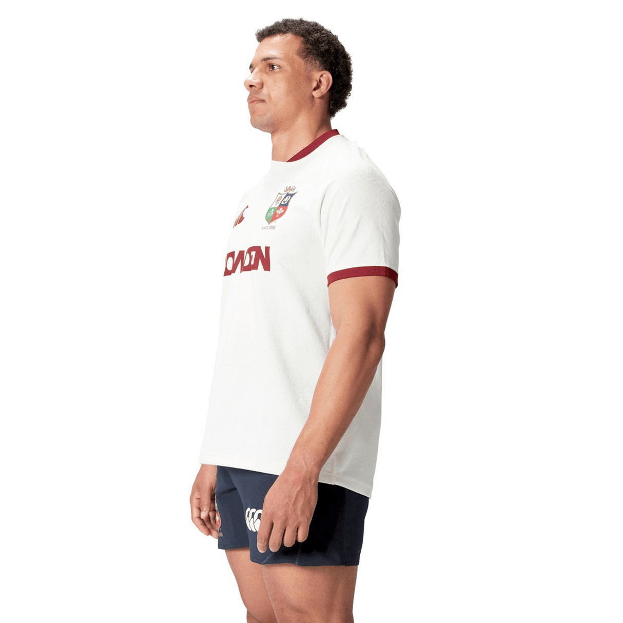 A man wearing the British & Irish Lions 2025 Training Jersey by Canterbury, made from recycled polyester, pairs it with navy shorts as he stands sideways, looking ahead.