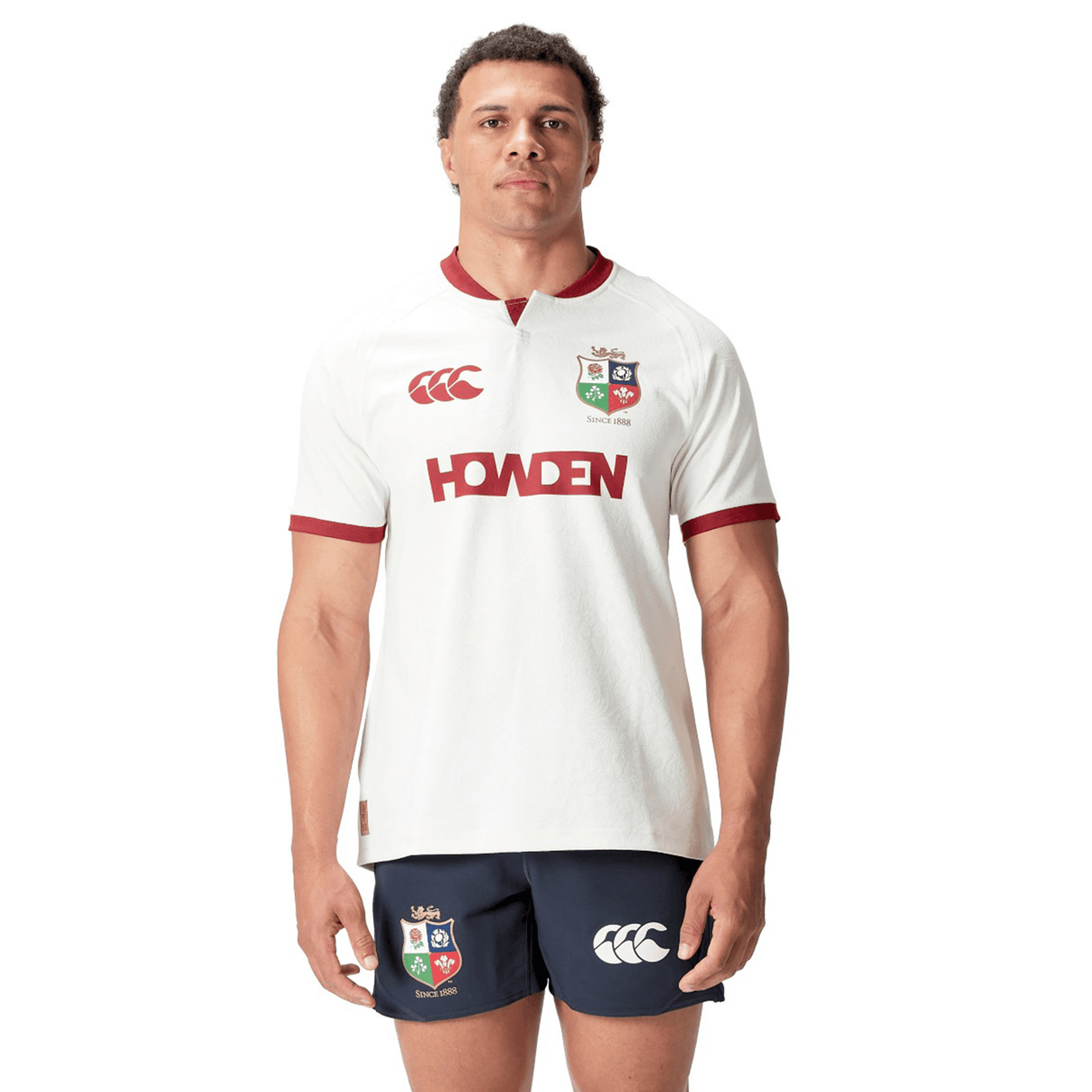 A person stands facing forward wearing a white British & Irish Lions 2025 Training Jersey by Canterbury, made from recycled polyester, paired with navy shorts featuring logos.