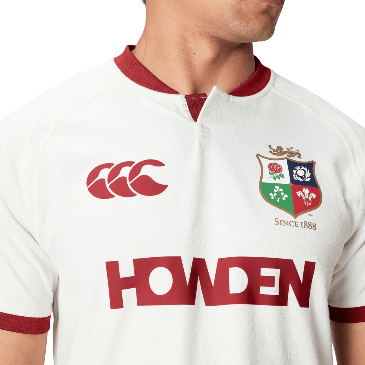 A person wearing the British & Irish Lions 2025 Training Jersey by Canterbury, a white rugby jersey with red accents, featuring "HOWDEN," a logo with three feathers, and "SINCE 1888," showcasing ergonomically placed panels inspired by the iconic British & Irish Lions design.