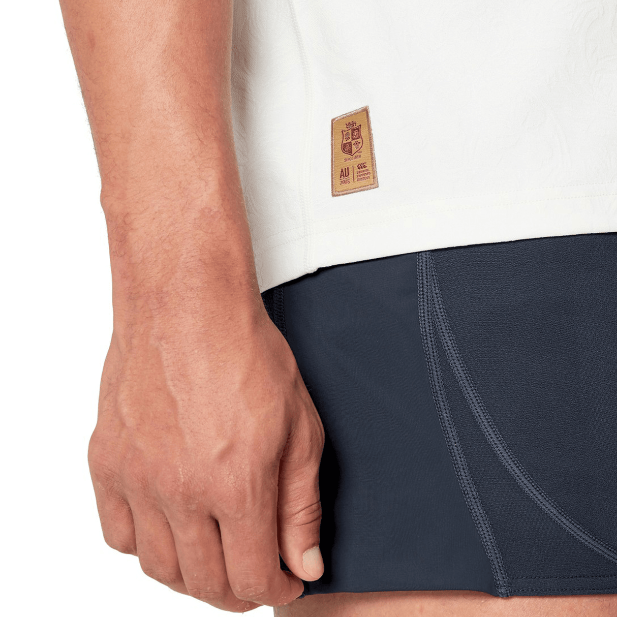 Close-up of a person’s hand resting by their side, wearing a white top with Canterbury branding and dark athletic shorts featuring visible stitching. A small label on the top is reminiscent of the British & Irish Lions 2025 Training Jersey by Canterbury, designed with ergonomically placed panels for comfort.