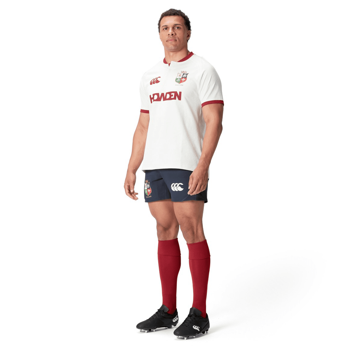 A rugby player wearing the British & Irish Lions 2025 Training Jersey by Canterbury, composed of recycled polyester and featuring a white and red design, stands against a white backdrop, glancing to the side.