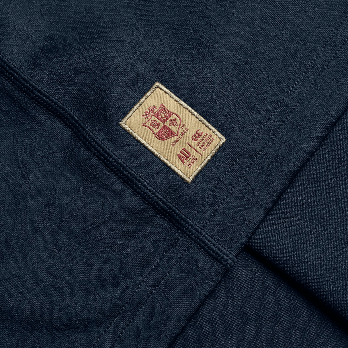 Crafted by Canterbury, the British & Irish Lions 2025 Training Jersey showcases a dark fabric adorned with a beige patch. This patch features a small crest design and text, akin to the iconic replica training jersey. Seamlessly sewn onto the smooth material, it perfectly integrates with strategically placed panels for enhanced comfort.
