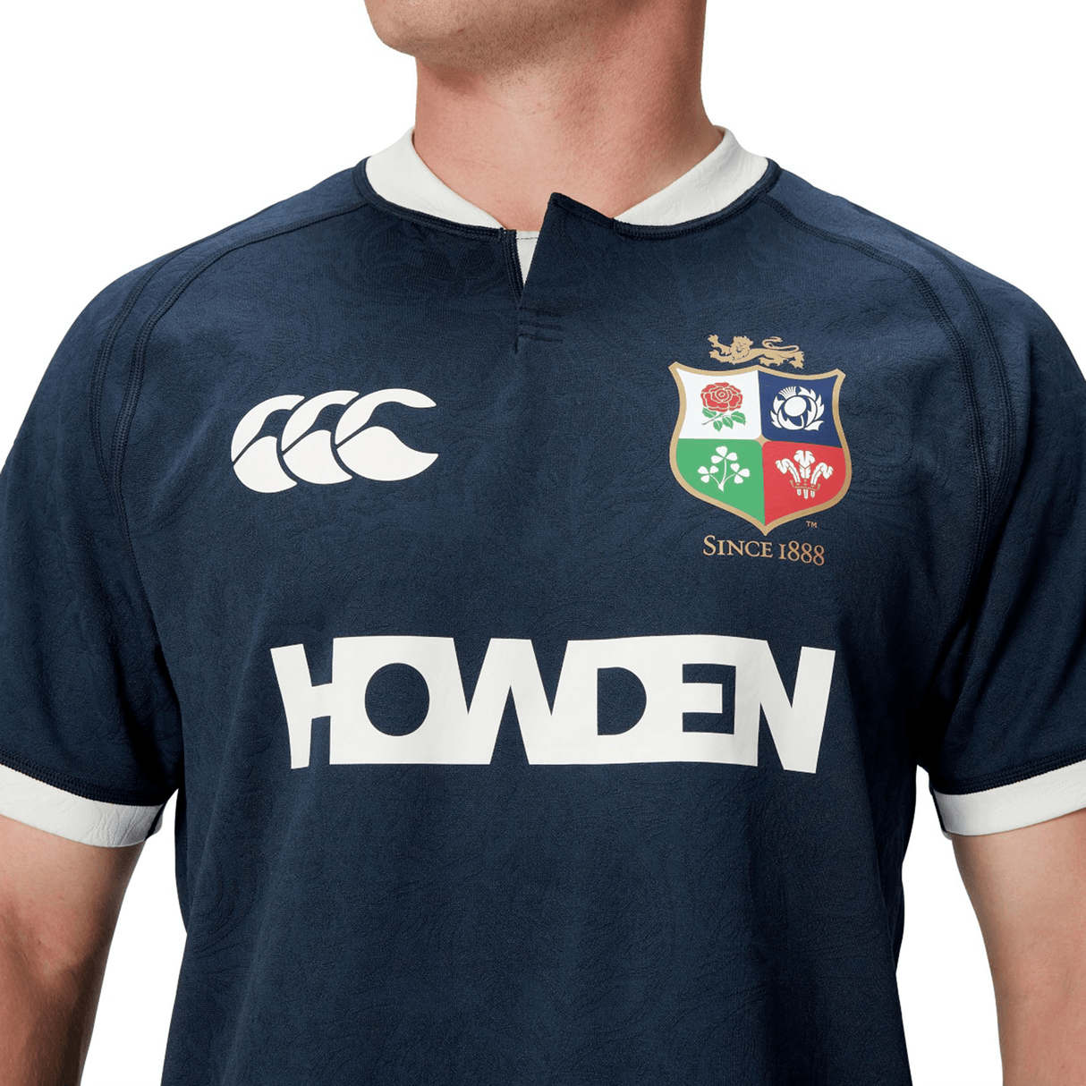 A person wearing the British & Irish Lions 2025 Training Jersey by Canterbury, featuring a navy blue design with logos and the text "Howden" on the front, along with ergonomically placed panels for enhanced comfort.