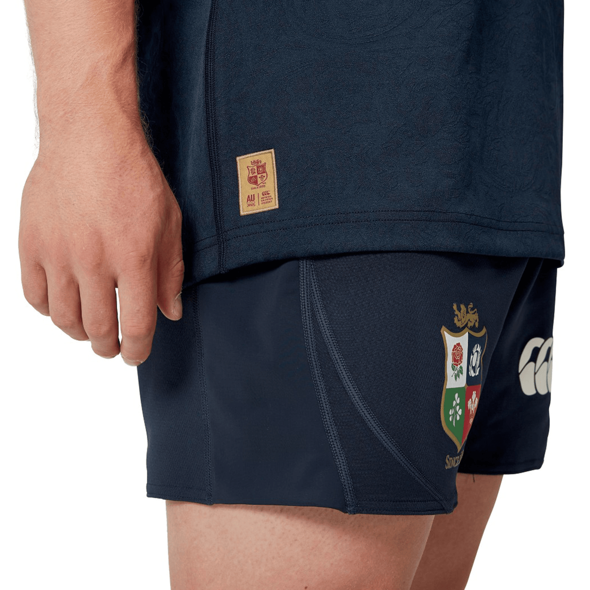 A person in a dark sports outfit is wearing the British & Irish Lions 2025 Training Jersey by Canterbury, which features ergonomically placed panels, along with shorts embellished with the colorful crest and logo of the British & Irish Lions.