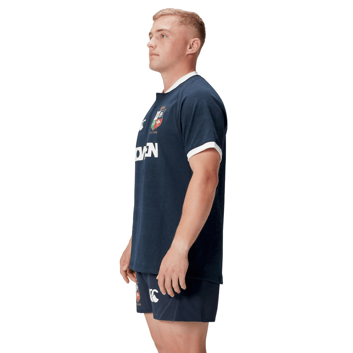 A man stands in profile wearing the British & Irish Lions 2025 Training Jersey by Canterbury. This navy rugby uniform is adorned with red and white crest logos, inspired by the classic British & Irish Lions design, and crafted from recycled polyester, combining style and sustainability.