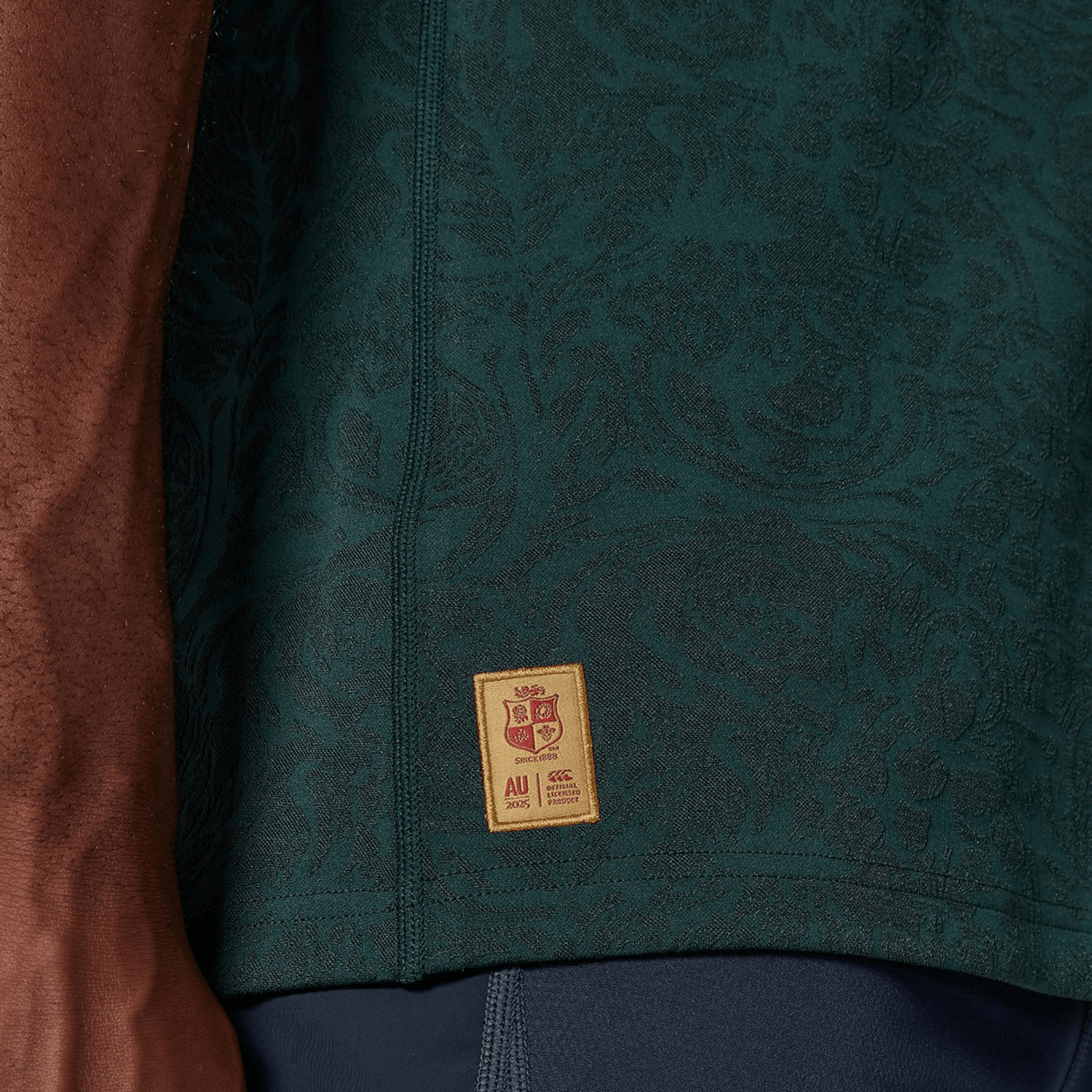 Close-up of a person wearing the British & Irish Lions 2025 Training Jersey by Canterbury, showcasing a dark green pattern with a small yellow and red rectangular patch at the bottom.