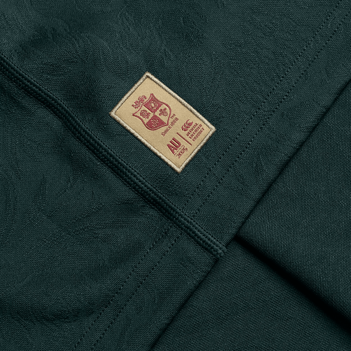 Close-up of a dark green fabric, crafted from recycled polyester, adorned with a beige patch stitched with a maroon crest and the year 2014 alongside the letters "AU," evoking the iconic style of Canterbury's British & Irish Lions 2025 Training Jersey.