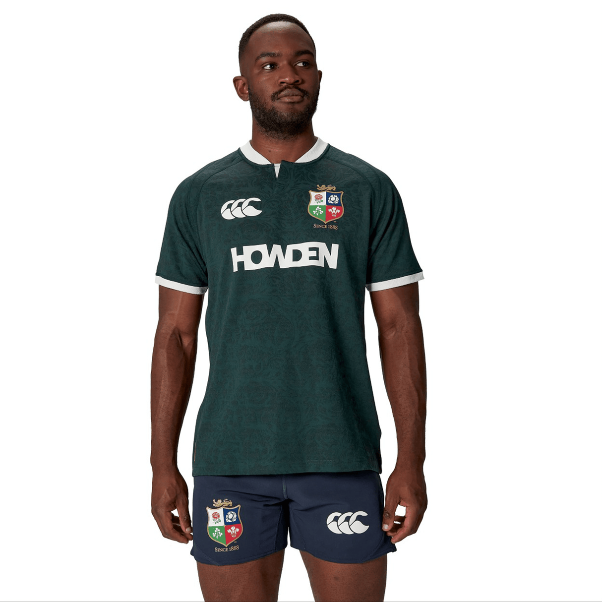 A man dressed in the British & Irish Lions 2025 Training Jersey by Canterbury, adorned with "Howden" and team logos, pairs it with navy shorts as he stands against a white background.