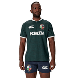A man dressed in the British & Irish Lions 2025 Training Jersey by Canterbury, adorned with "Howden" and team logos, pairs it with navy shorts as he stands against a white background.
