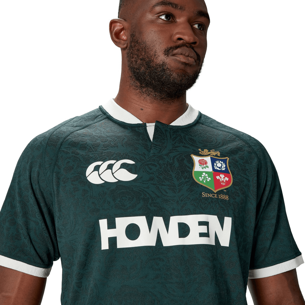 A man wearing the British & Irish Lions 2025 Training Jersey by Canterbury, featuring logos and "Howden" text. This stylish, dark green piece is crafted from recycled polyester for an eco-friendly touch.