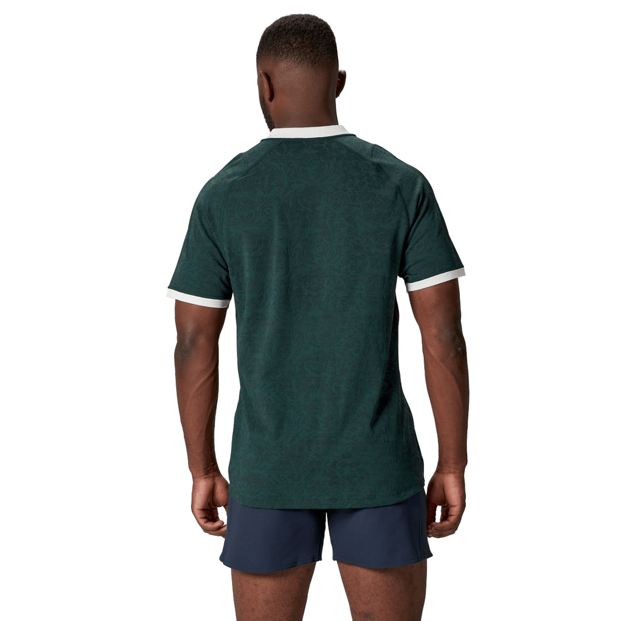 A person wearing the British & Irish Lions 2025 Training Jersey by Canterbury, made of recycled polyester in dark green, matched with dark shorts, stands facing away from the camera against a white background.