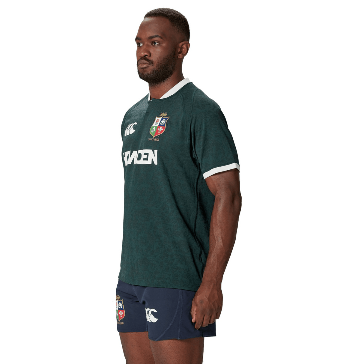 A man in a Canterbury-designed green and blue sports uniform, crafted from recycled polyester, stands facing sideways, closely resembling the British & Irish Lions 2025 Training Jersey.