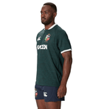 A man in a Canterbury-designed green and blue sports uniform, crafted from recycled polyester, stands facing sideways, closely resembling the British & Irish Lions 2025 Training Jersey.
