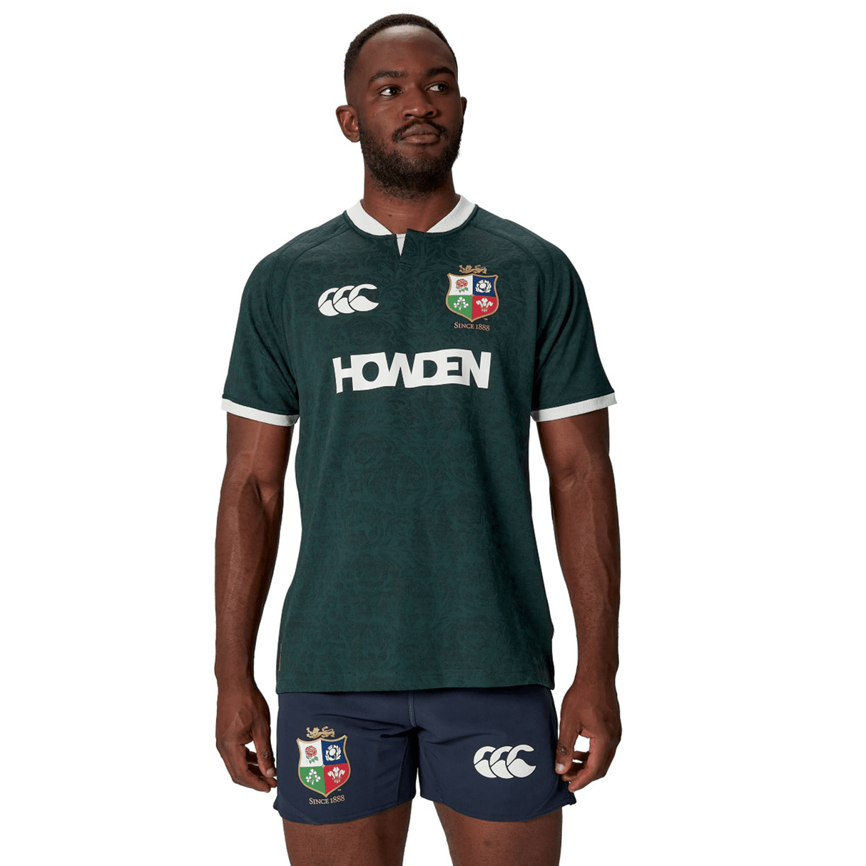 A man in a British & Irish Lions 2025 Training Jersey by Canterbury, crafted from recycled polyester and reminiscent of a dark green rugby jersey, paired with navy shorts, stands looking to the side.