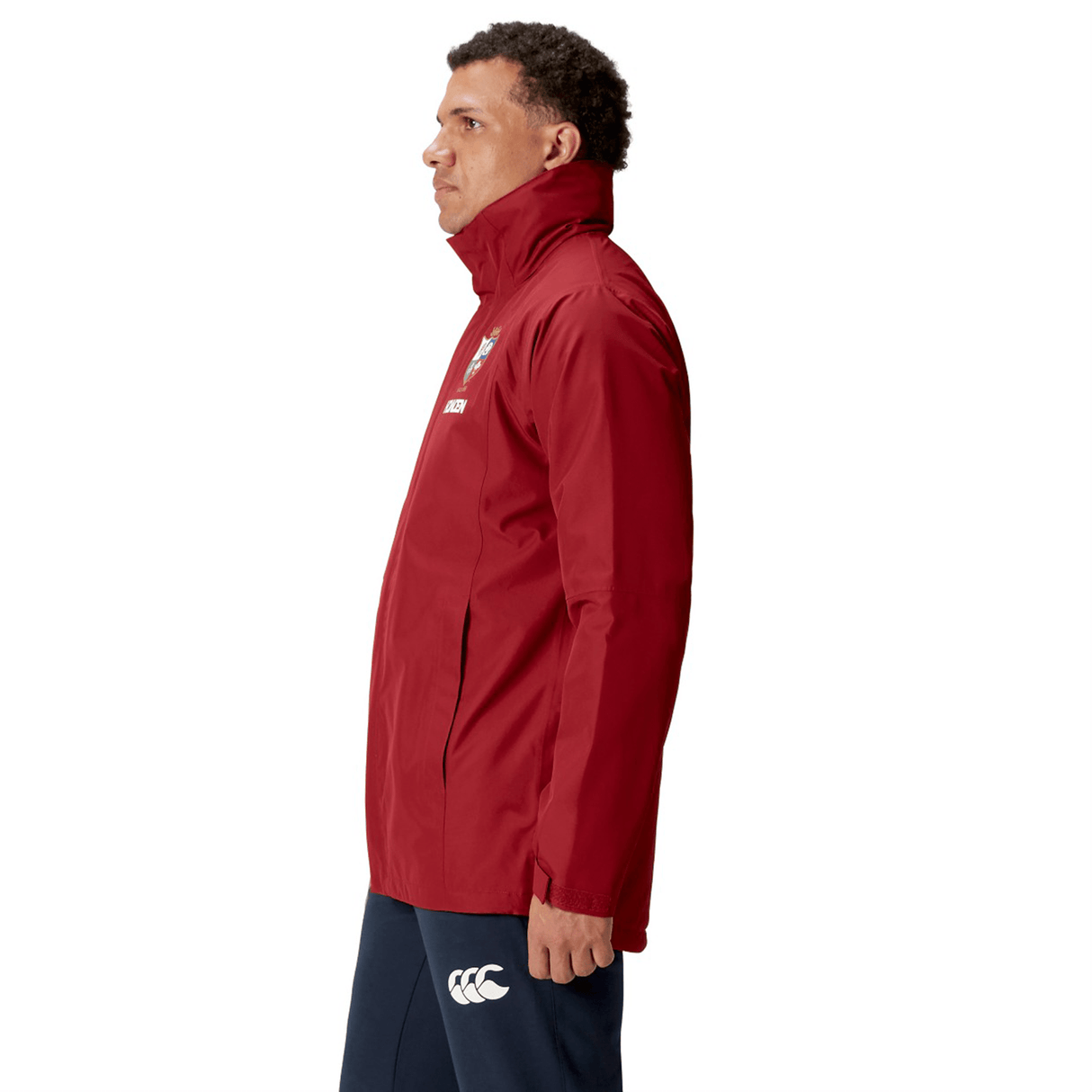 A person wearing a red British & Irish Lions 2025 Waterproof Jacket by Canterbury, featuring an insulated inner layer, and dark pants stands in profile against a white background.