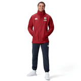 A person in a British & Irish Lions 2025 Waterproof Jacket by Canterbury, featuring logos, stands smiling with hands in pockets, wearing black pants and white sneakers against a white background.