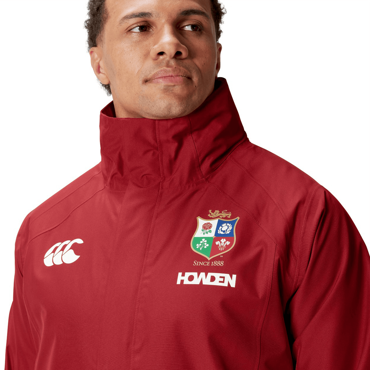 A person wearing the British & Irish Lions 2025 Waterproof Jacket by Canterbury, crafted from polyester in a red hue, showcasing the lion and crest logo of the British & Irish Lions with "SINCE 1888" text.
