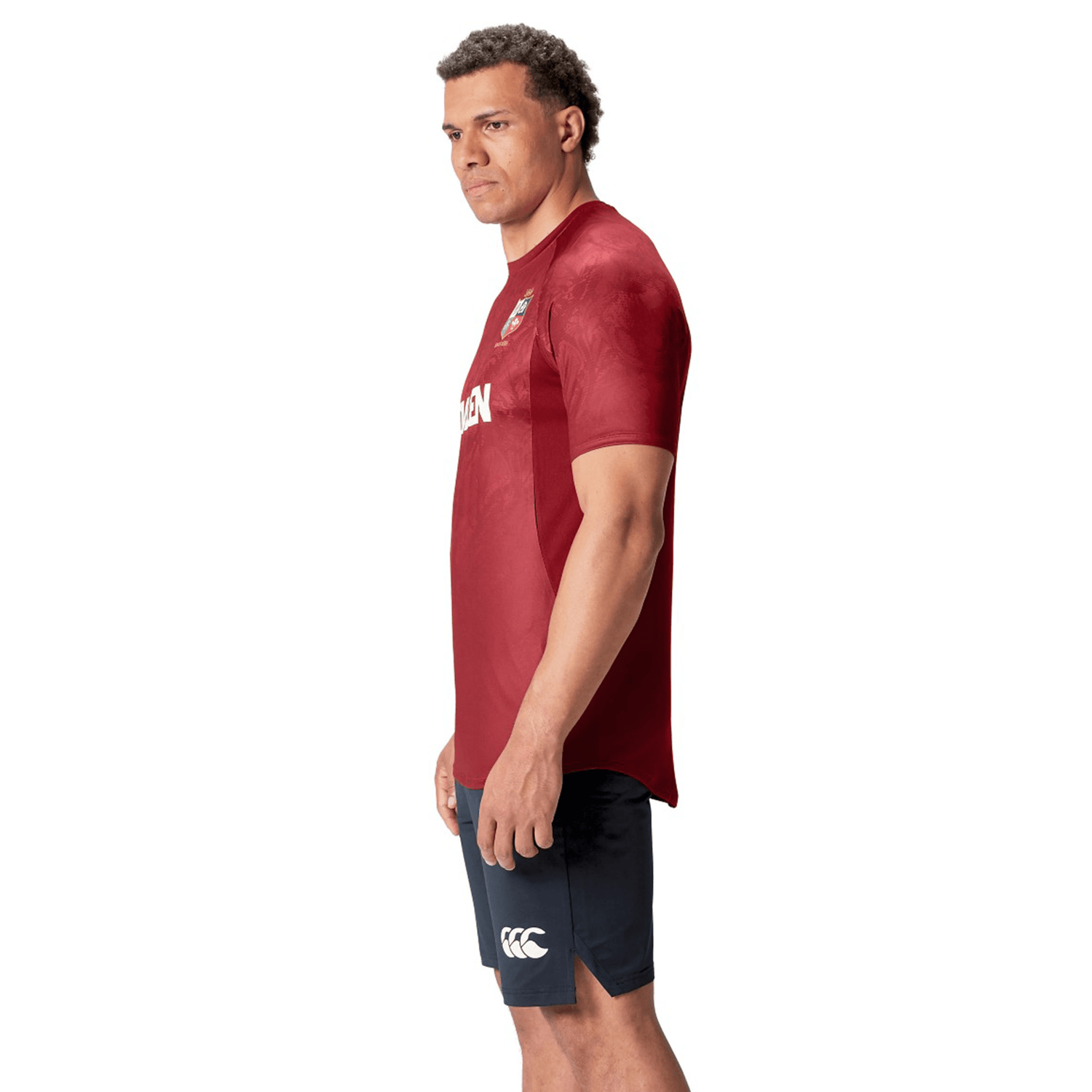 A man stands in a red British & Irish Lions 25 Superlight Training Tee by Canterbury, paired with navy shorts, embodying the iconic style of the British & Irish Lions with a neutral expression.