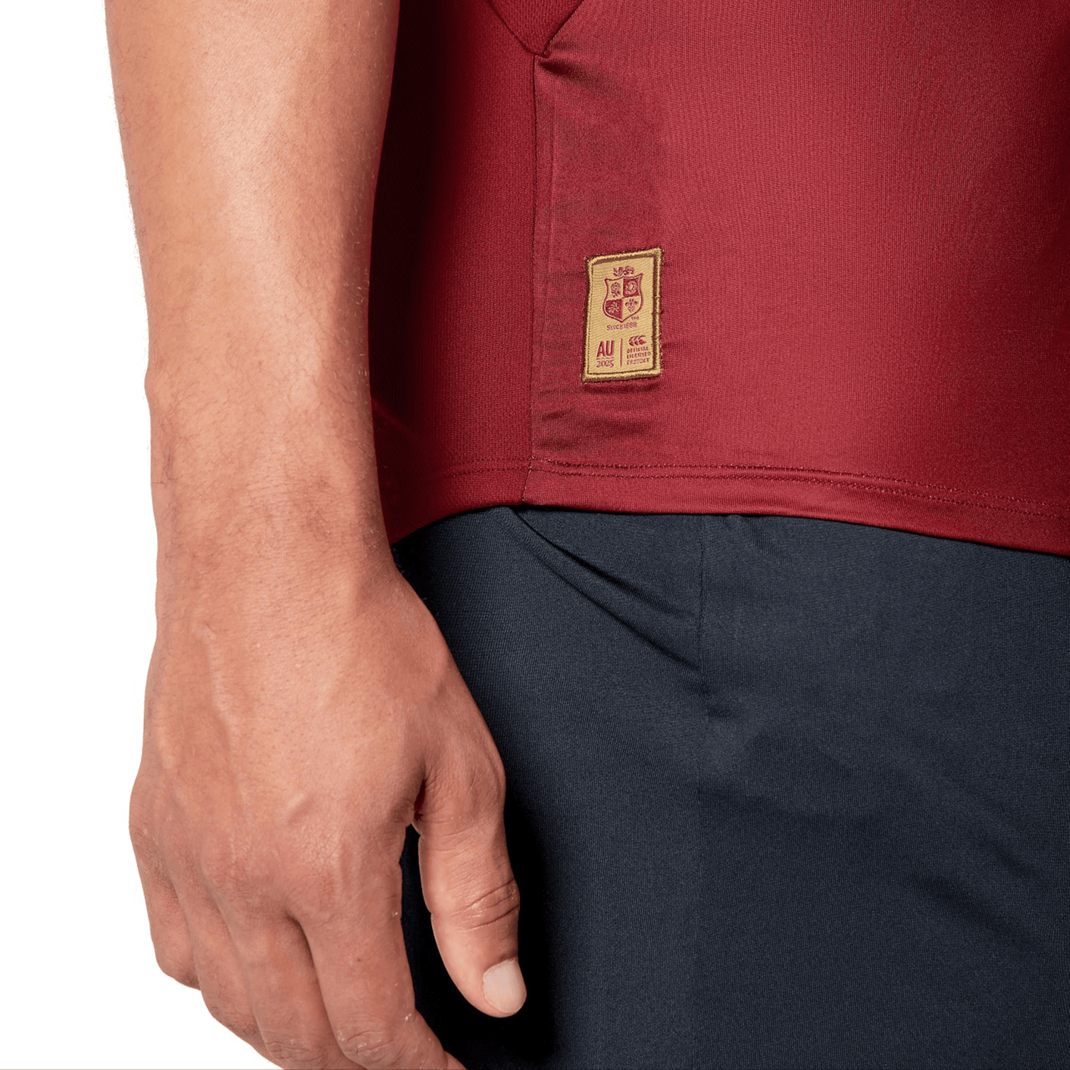 Close-up of a person wearing a red British & Irish Lions 25 Superlight Training Tee by Canterbury paired with dark pants. The tee includes a beige patch featuring a crest reminiscent of the British & Irish Lions and small text near the hem. The person's left arm rests calmly by their side.