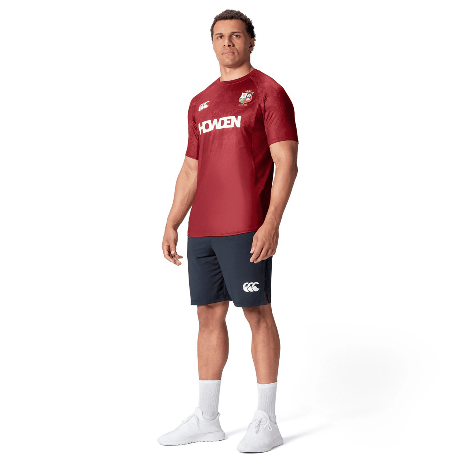 A person stands wearing a British & Irish Lions 25 Superlight Training Tee by Canterbury, featuring "HOWDEN" across the chest, paired with navy shorts, white socks, and white sneakers.