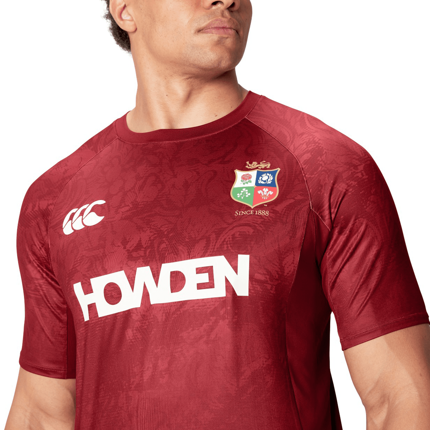 A man wears a red rugby jersey prominently displaying the "HOWDEN" name and team logo, reminiscent of the British & Irish Lions' style. This design is inspired by the British & Irish Lions 25 Superlight Training Tee by Canterbury, offering fans a perfect blend of comfort and heritage.
