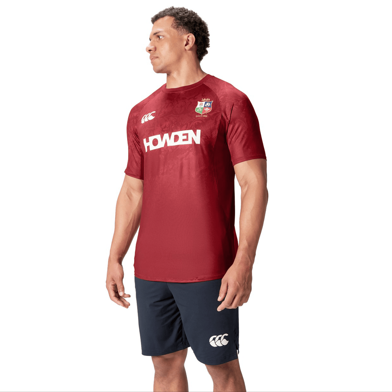 A man wearing a red British & Irish Lions 25 Superlight Training Tee by Canterbury, featuring the word "Howden," stands against a white background.