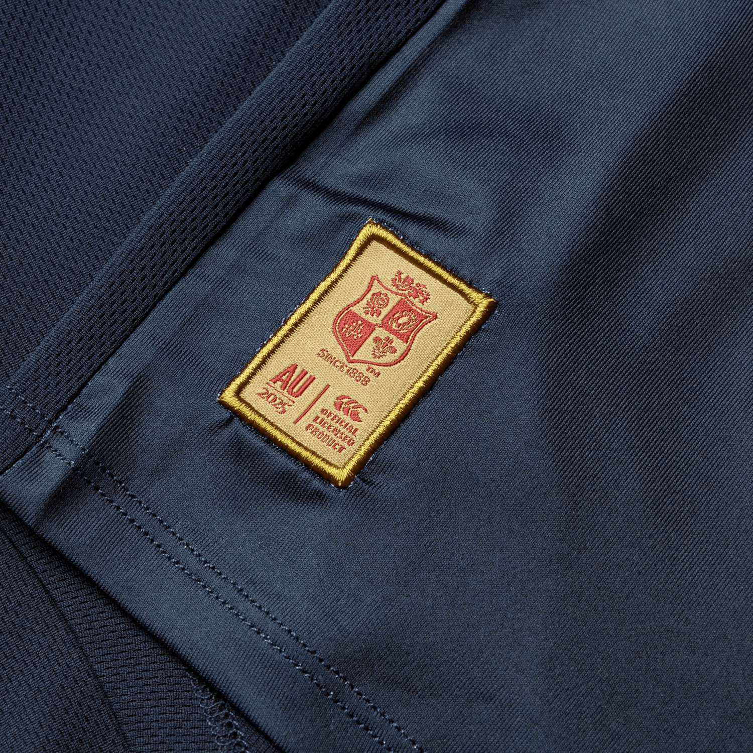 Close-up of a dark blue fabric on the British & Irish Lions 25 Superlight Training Tee by Canterbury, featuring a rectangular gold patch with a red shield, "since 1888," "AU 2025," and other text. The patch boasts a meticulously sewn border.