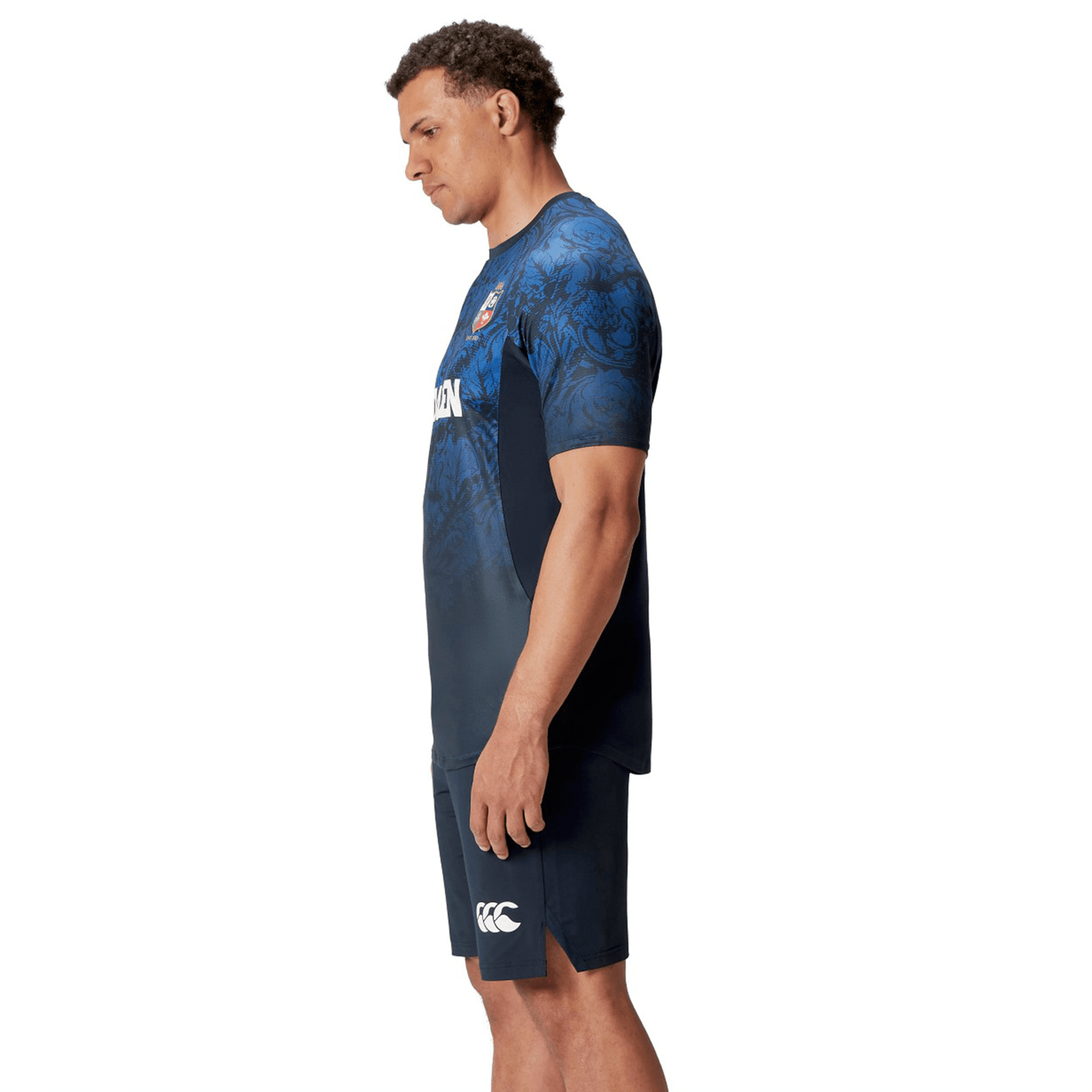 A person wearing a British & Irish Lions 25 Superlight Training Tee by Canterbury, featuring a blue and black floral pattern, stands with their hands by their sides, viewed from the side.