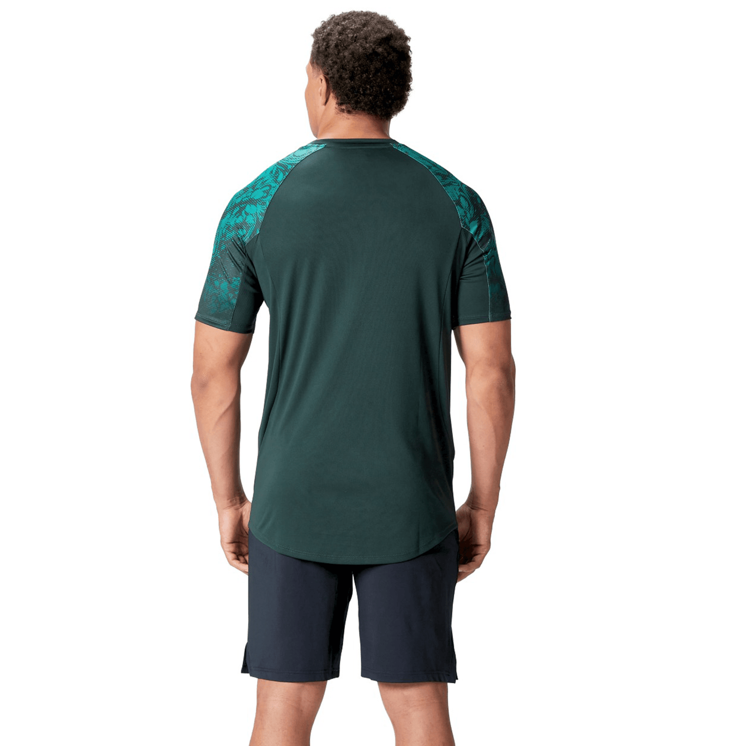A man stands with his back to the camera, wearing a dark green British & Irish Lions 25 Superlight Training Tee by Canterbury with patterned sleeves and black shorts.