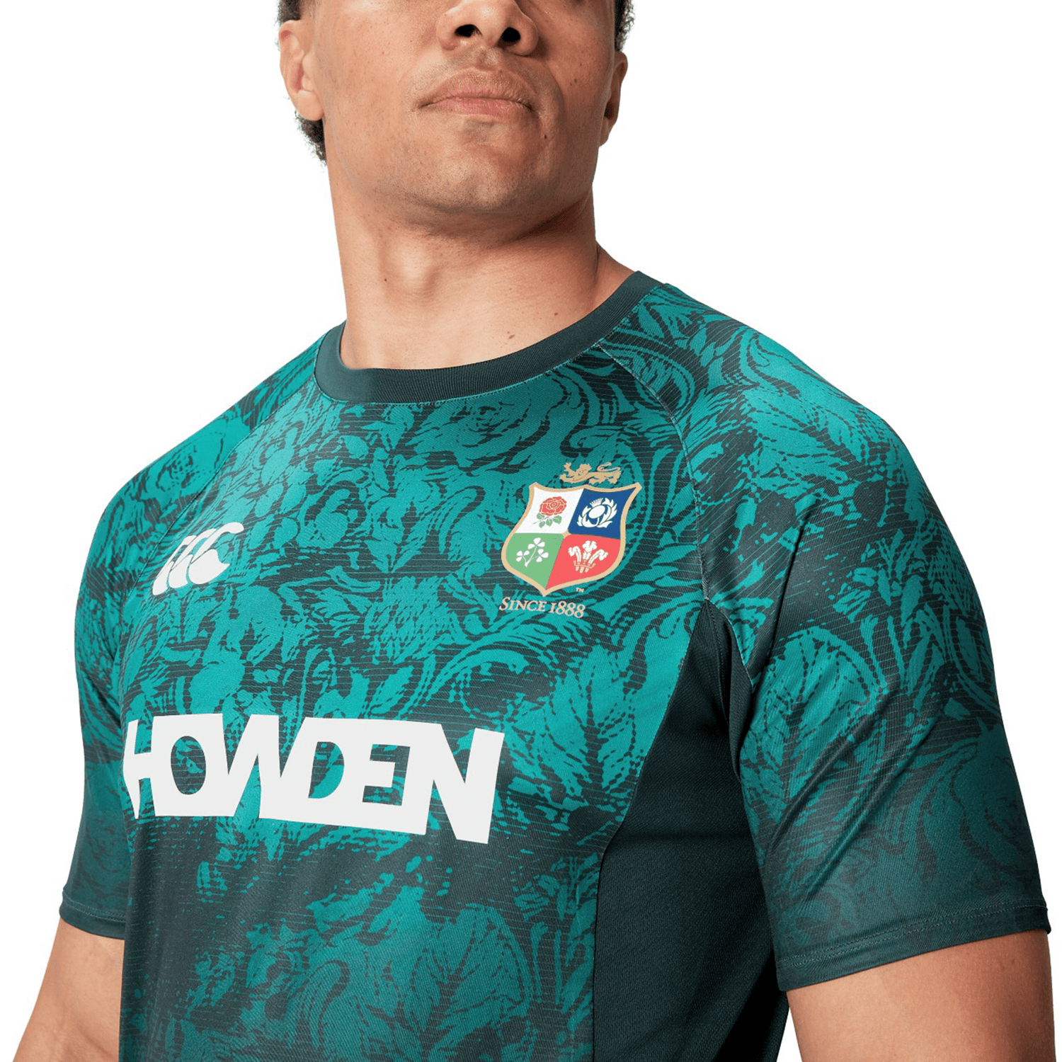 A person wearing the British & Irish Lions 25 Superlight Training Tee by Canterbury, featuring a green rugby jersey with a patterned design and logos, including "Howden" and a crest badge, reminiscent of the iconic style by Canterbury.