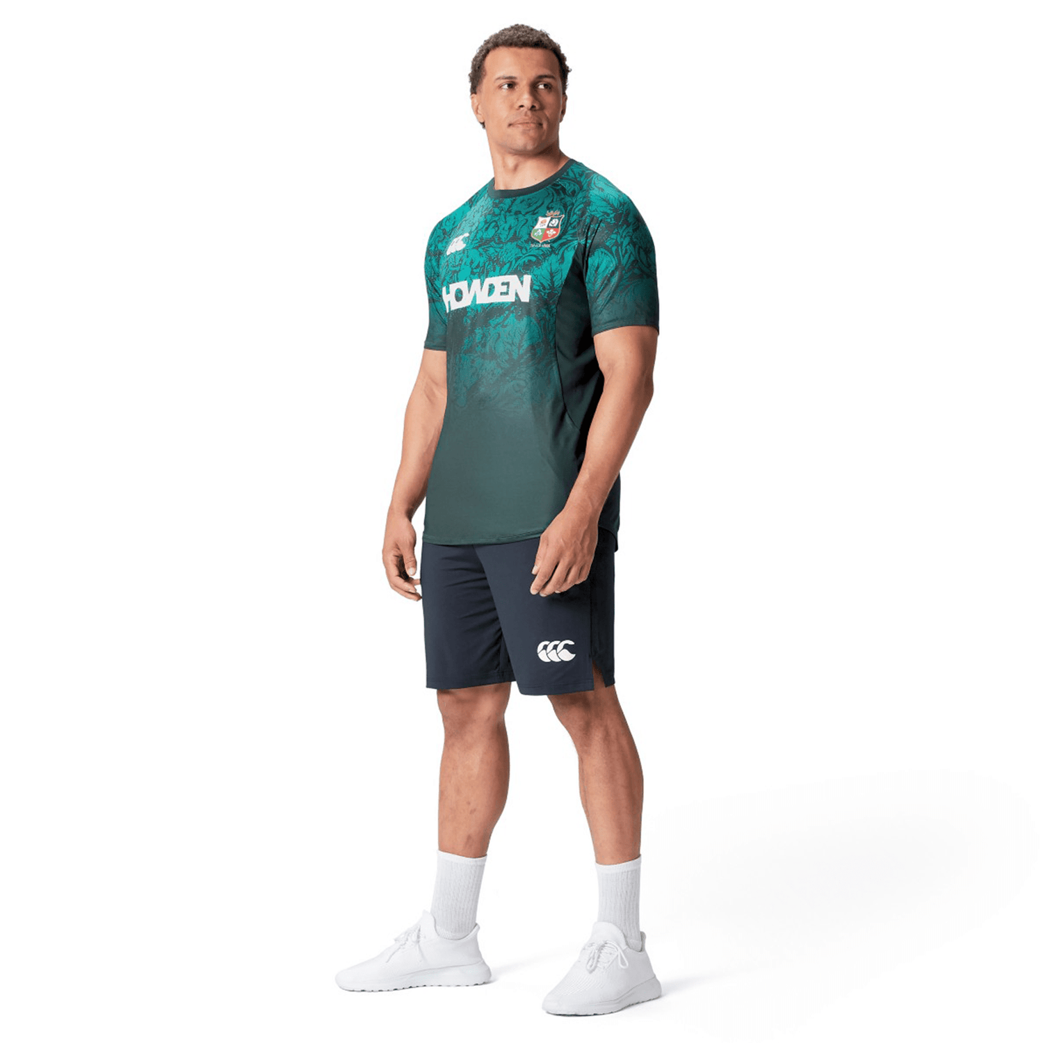 A man wearing a green and blue jersey, reminiscent of the British & Irish Lions 25 Superlight Training Tee by Canterbury, and navy shorts stands facing slightly to the left. He is sporting white socks and shoes against a plain white background.