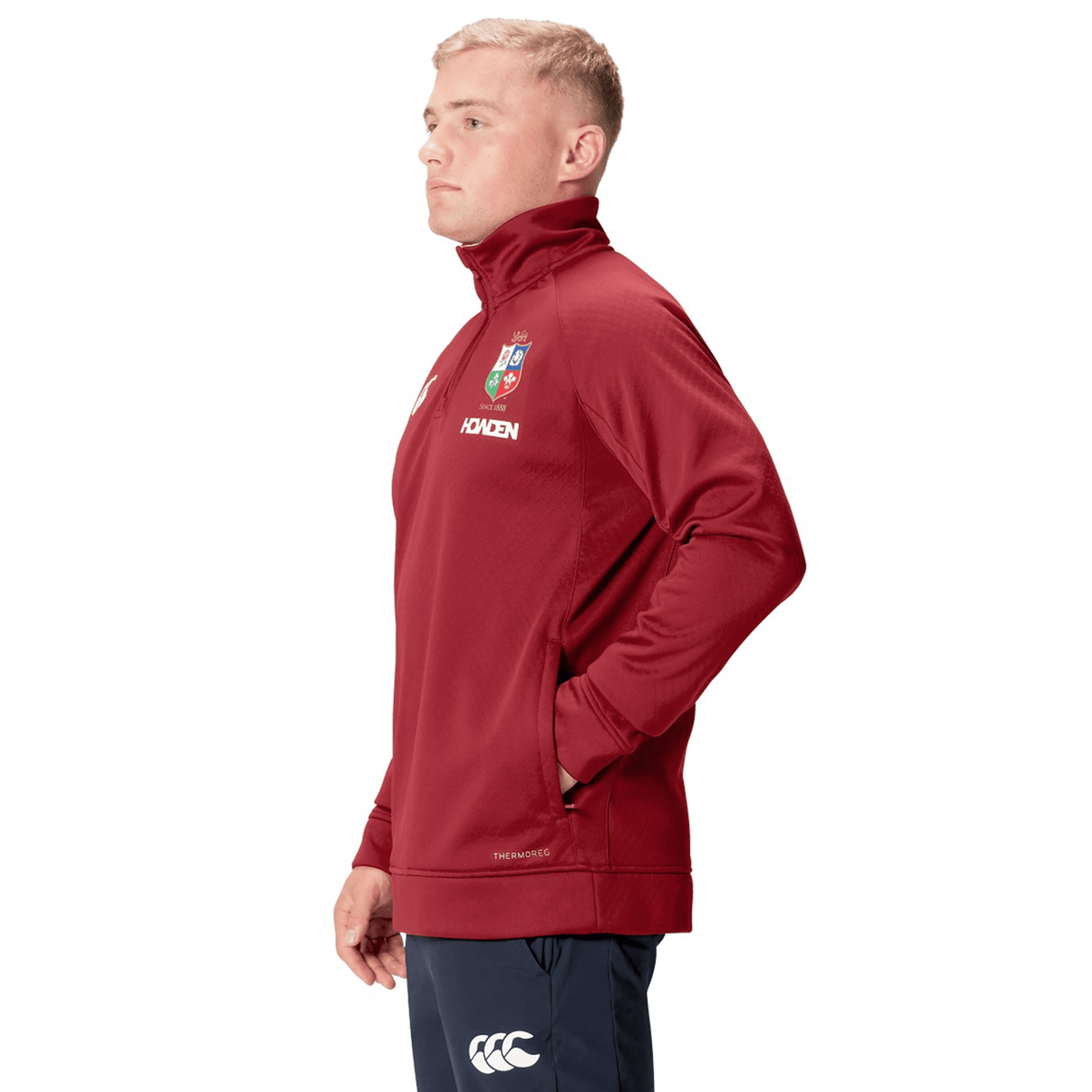 A person in the British & Irish Lions 2025 1/4 Zip Fleece by Canterbury stands sideways with hands in pockets, proudly showcasing their affiliation with the British & Irish Lions.
