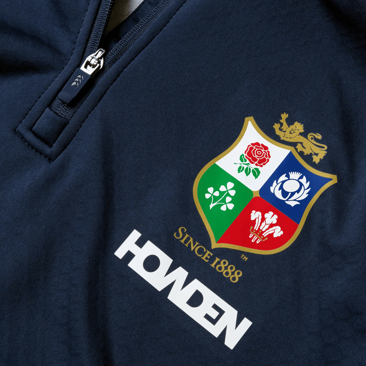 Close-up of a navy blue sports jersey featuring a vibrant shield logo with a lion, rose, shamrock, and thistle. The words "SINCE 1888" and "HOWDEN" are displayed below the emblem. Designed by Canterbury for the British & Irish Lions, this item pairs perfectly with their British & Irish Lions 2025 1/4 Zip Fleece by Canterbury.