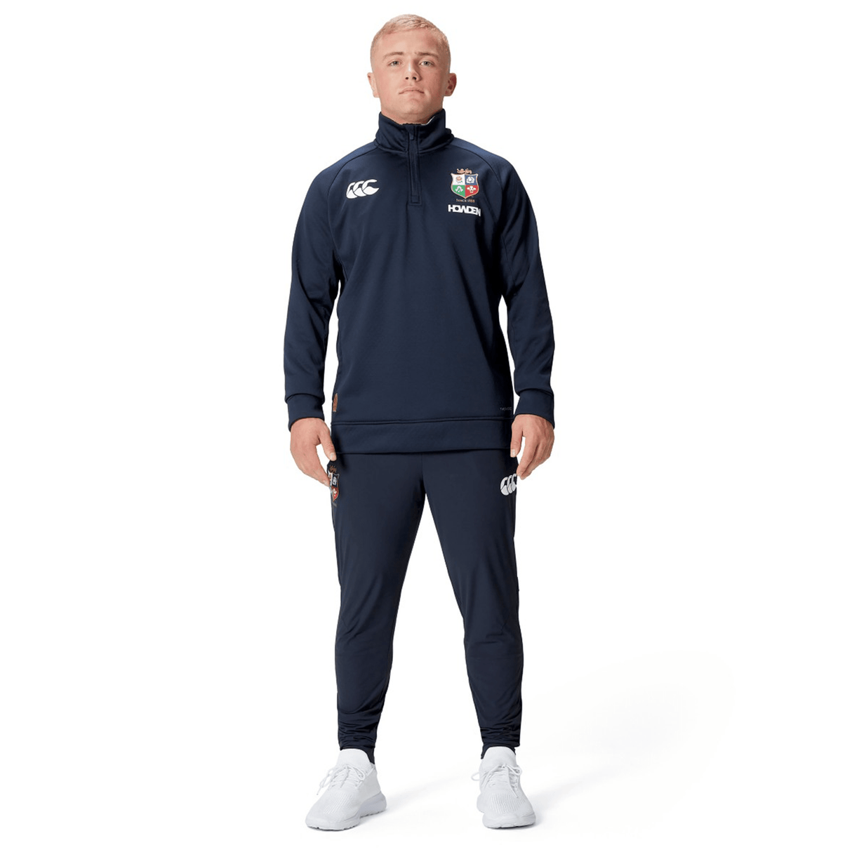 A person dressed in a navy British & Irish Lions 2025 1/4 Zip Fleece by Canterbury and white shoes stands against a clean white background, exuding the timeless elegance associated with the British & Irish Lions.