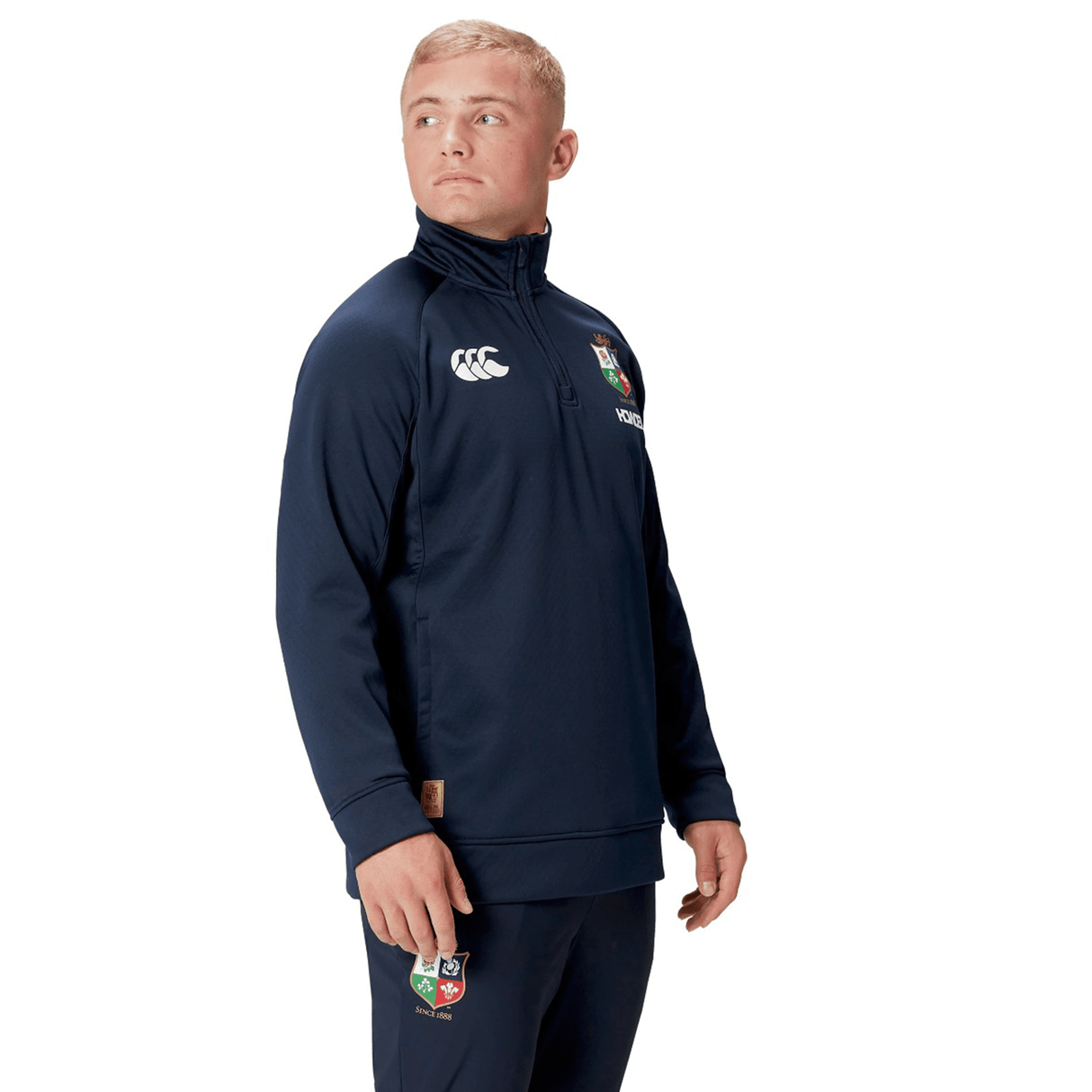 A person clad in the British & Irish Lions 2025 1/4 Zip Fleece by Canterbury, featuring a logo on the chest and pants, stands against a white background.