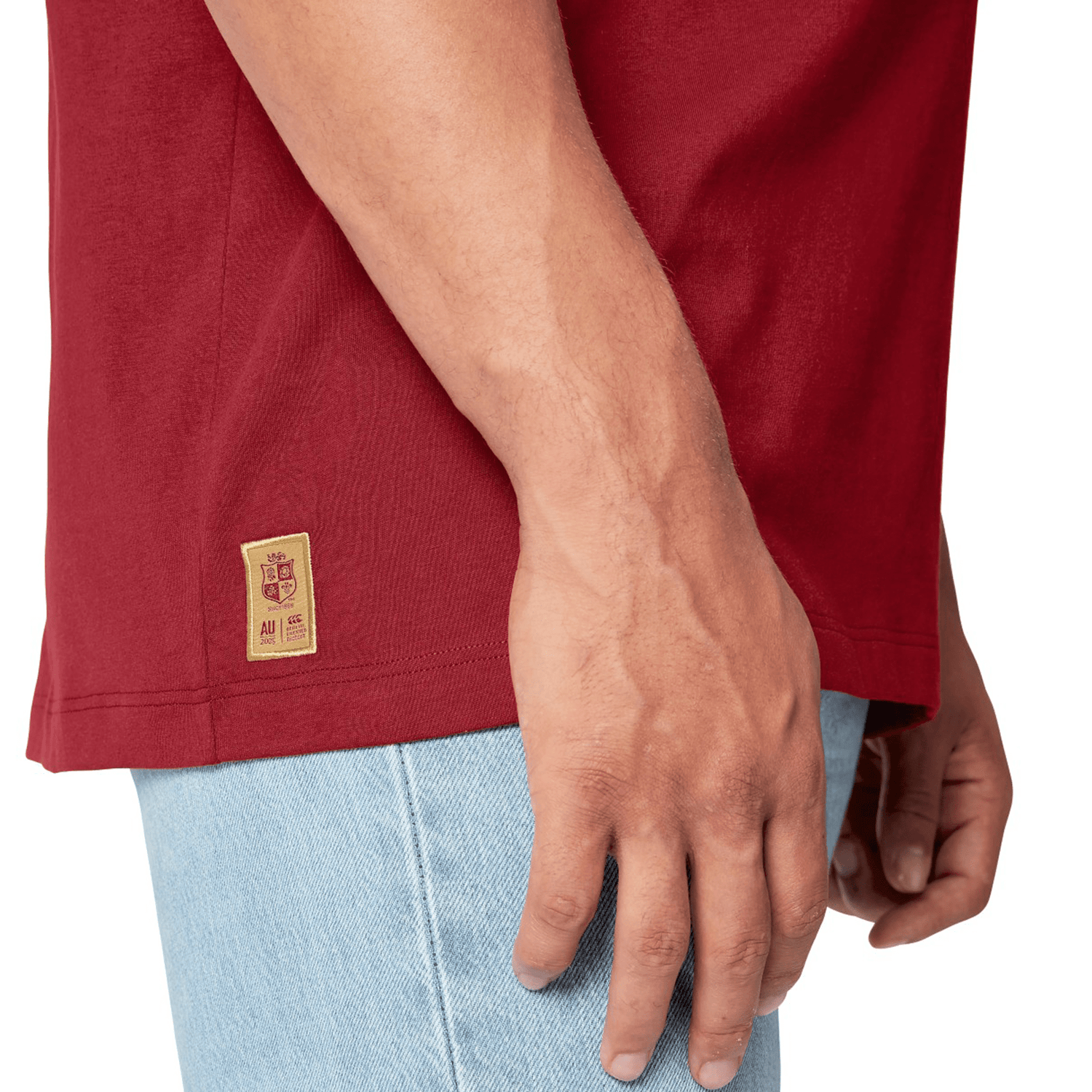 A person wearing a British & Irish Lions 2025 Cotton Jersey Tee by Canterbury, made of organic cotton, in red along with light blue jeans. The tee resembles those worn by rugby supporters and features a small patch near the hem on the left side.