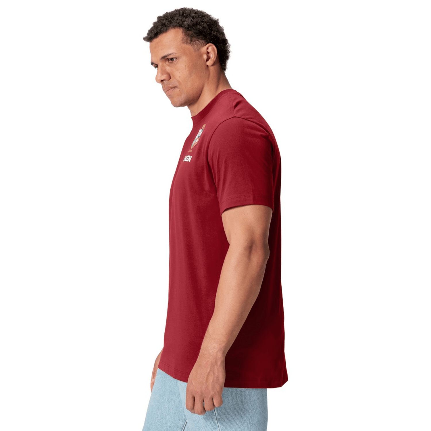 A man wearing the British & Irish Lions 2025 Cotton Jersey Tee by Canterbury and light blue jeans stands against a white background, facing slightly left. He has the confident stance of a rugby supporter, reminiscent of fans cheering for the British & Irish Lions.