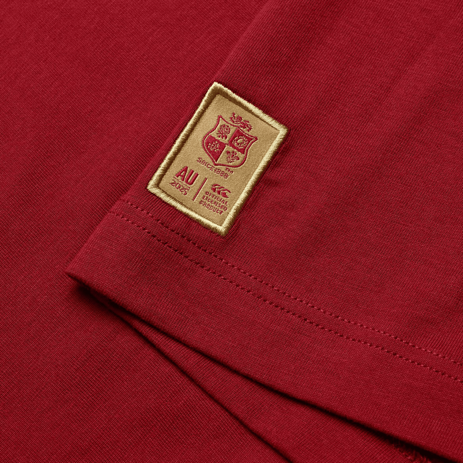 A close-up of the British & Irish Lions 2025 Cotton Jersey Tee by Canterbury showcases a stitched patch on red fabric, featuring a coat of arms, the letters "AU," and the year "2025." Crafted from organic cotton, this tee is ideal for any rugby supporter, embodying the enduring spirit of the British & Irish Lions.