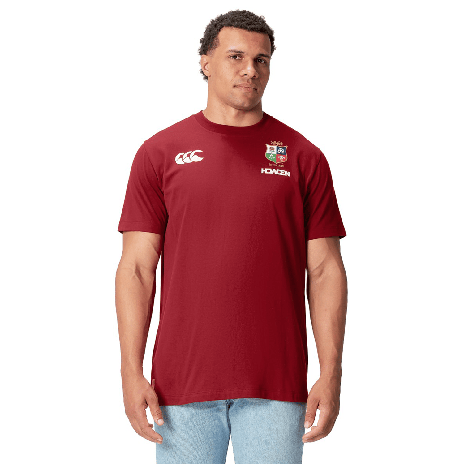 A rugby supporter proudly wears a British & Irish Lions 2025 Cotton Jersey Tee by Canterbury, featuring maroon and white logos, crafted from organic cotton, standing against a pristine white background.