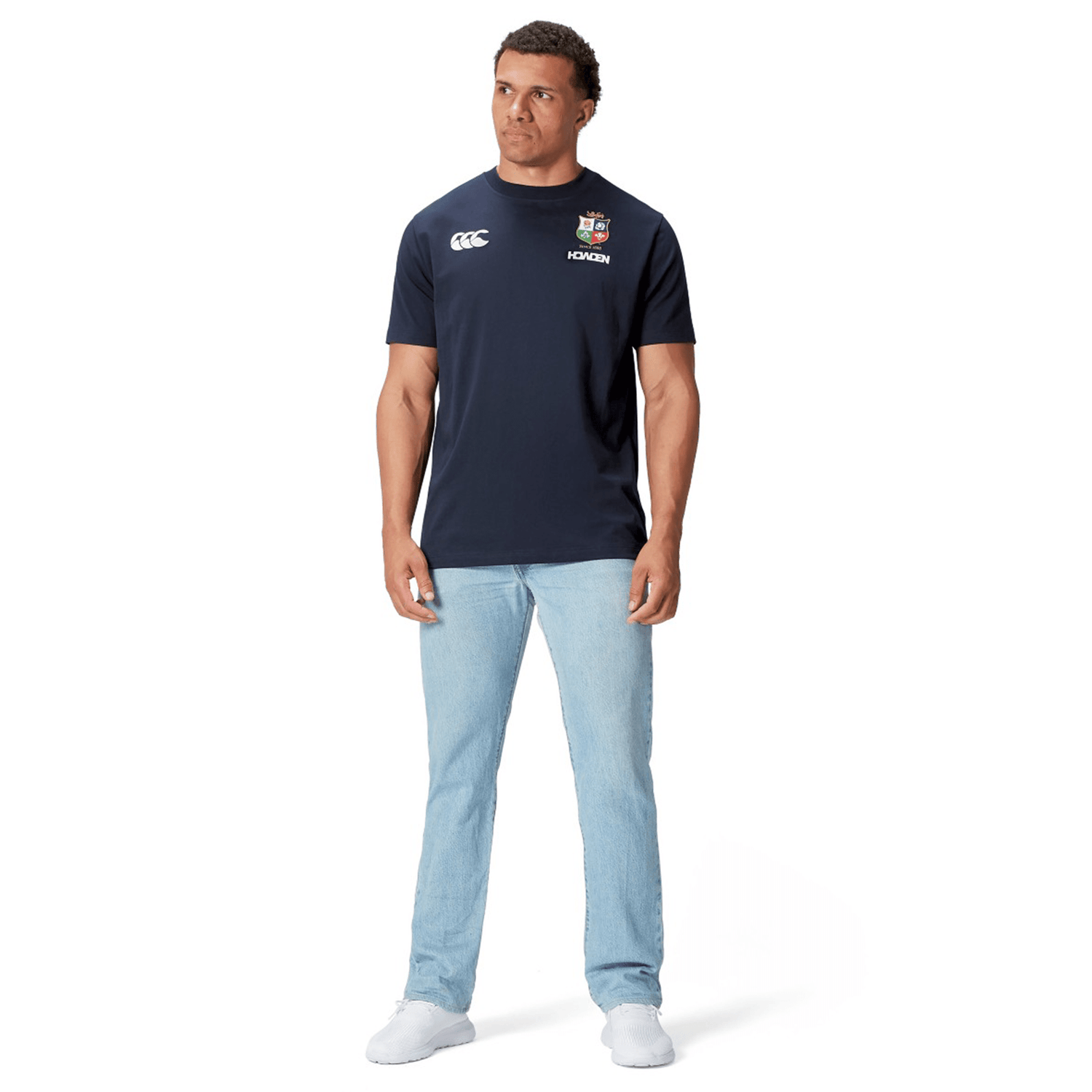 A rugby fan stands against a crisp white background, stylishly dressed in light blue jeans paired with the British & Irish Lions 2025 Cotton Jersey Tee by Canterbury, featuring logos and crafted from organic cotton.
