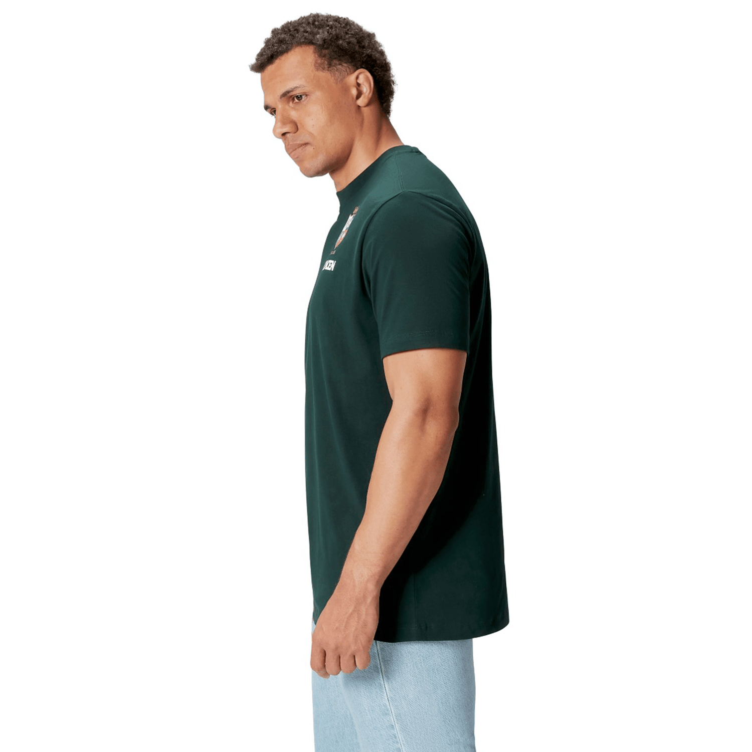 A rugby supporter wearing the British & Irish Lions 2025 Cotton Jersey Tee by Canterbury in a dark green shade, paired with light blue jeans, is viewed from the side against a plain white background.