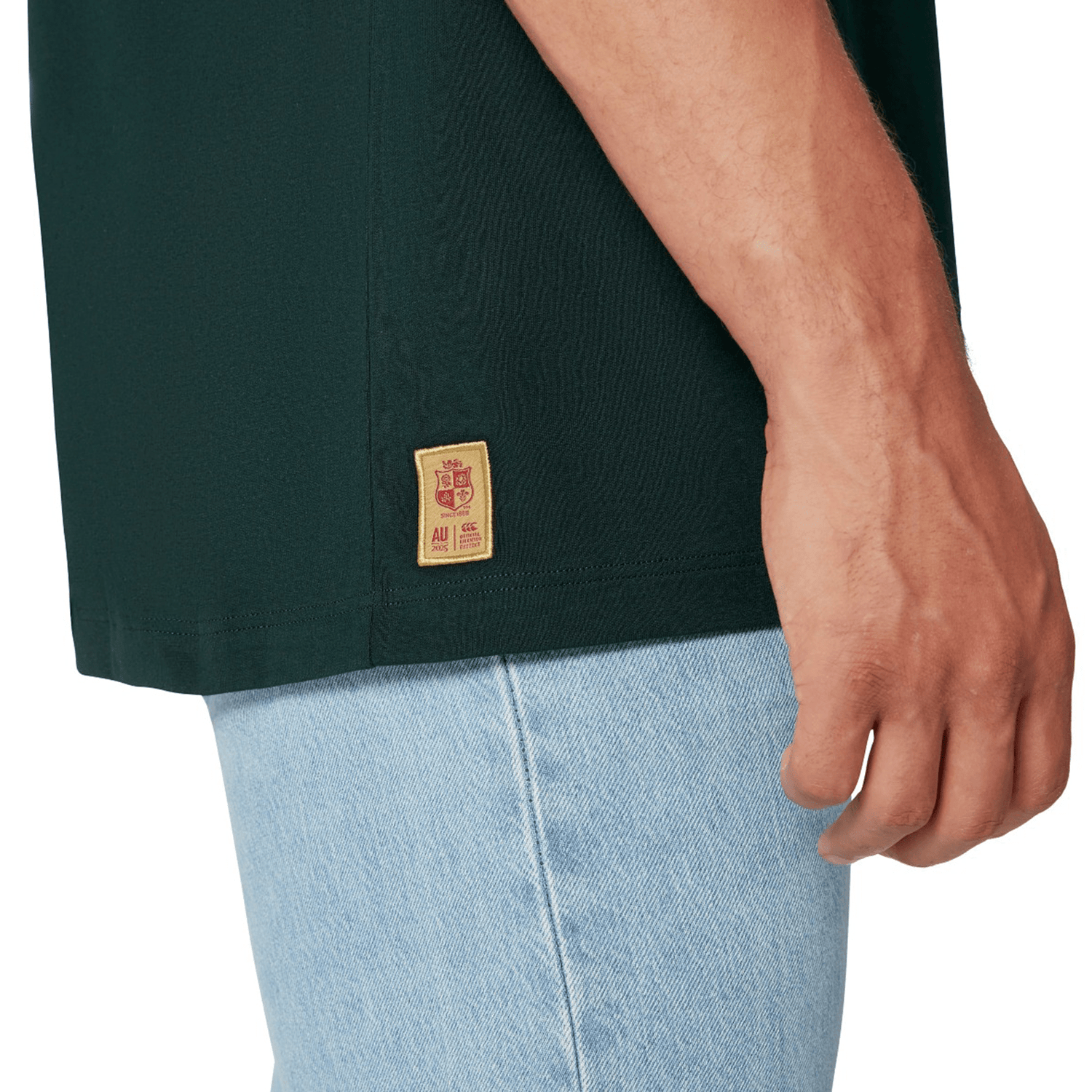 A person wearing the British & Irish Lions 2025 Cotton Jersey Tee by Canterbury, crafted from organic cotton in dark green with a discreet tag, paired with light blue jeans. The focus is on the torso and right hand, exuding the casual yet spirited vibe of a rugby supporter.