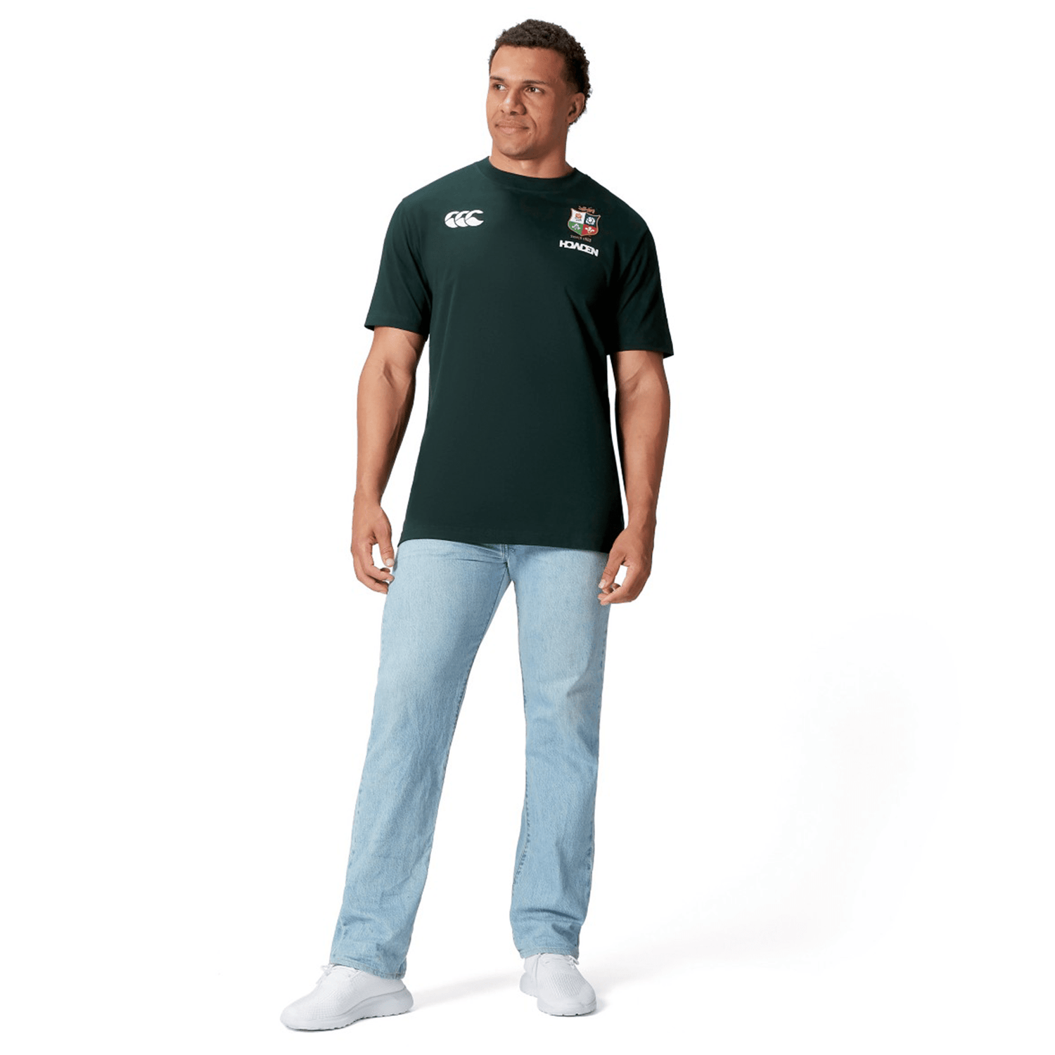 A rugby supporter wearing a dark green British & Irish Lions 2025 Cotton Jersey Tee by Canterbury, paired with light blue jeans and white sneakers, stands against a plain white background.