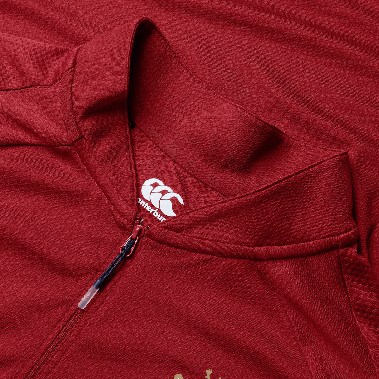 Close-up of a red British & Irish Lions 25 Everest First Layer Top by Canterbury, resembling a sports jersey, featuring a small white logo near the collar and a half-zipper partially opened.