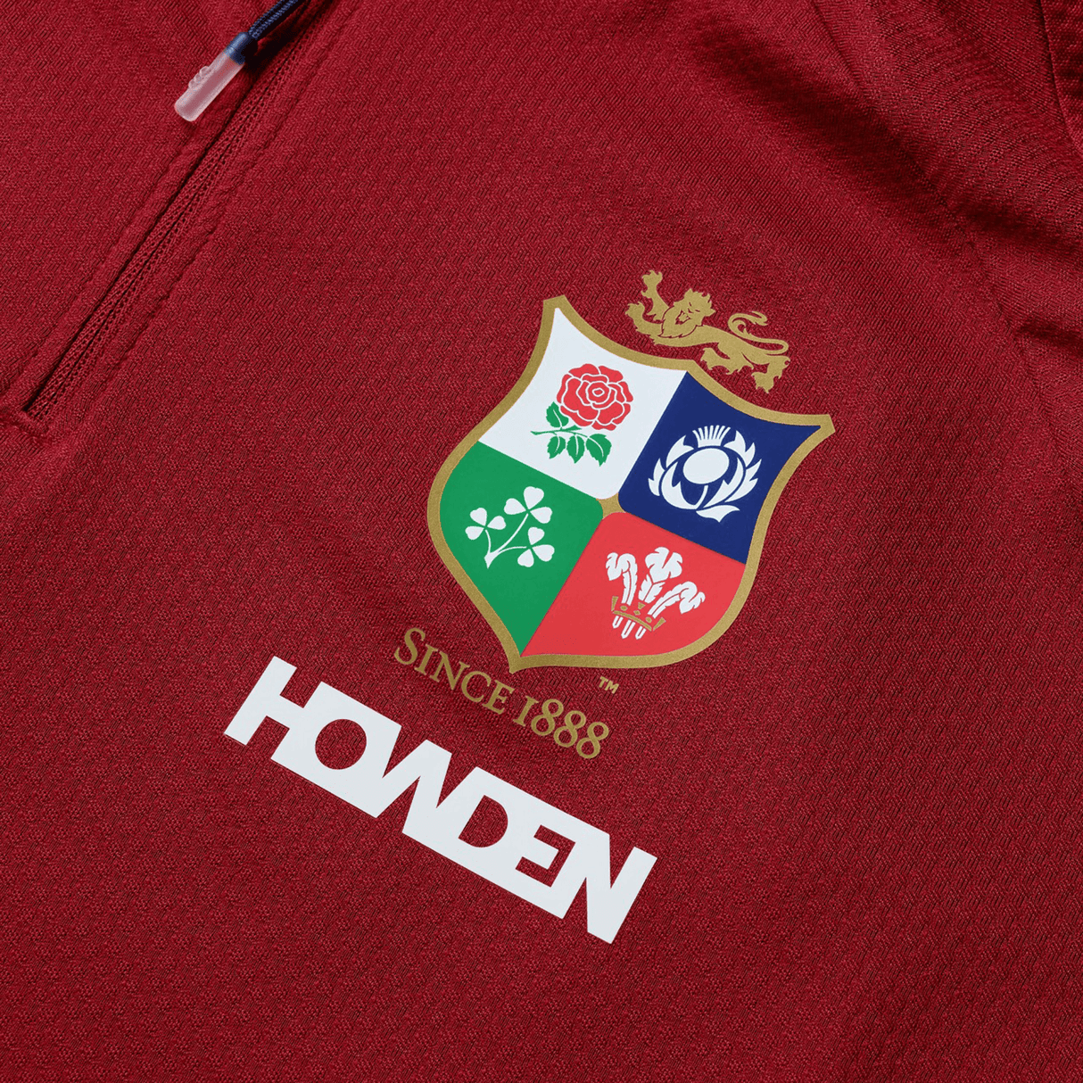 Canterbury's British & Irish Lions 25 Everest First Layer Top is a red sports jersey adorned with the iconic crest featuring a rose, thistle, shamrock, and dragon. The text "SINCE 1888" and "HOWDEN" are also displayed, making it perfect for passionate fans.