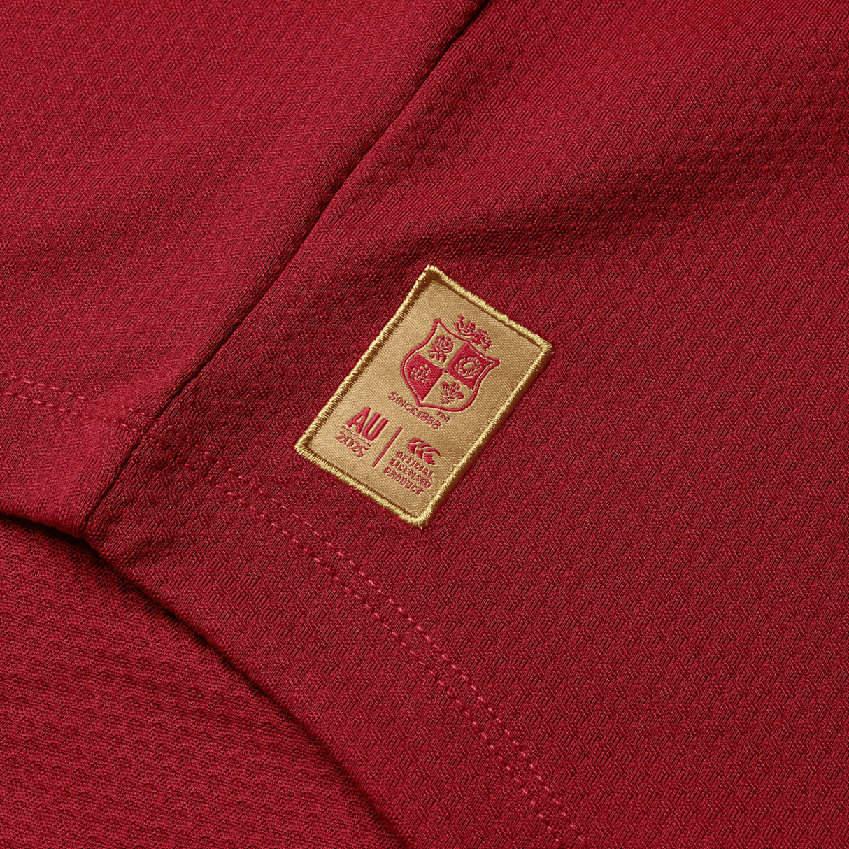 A close-up of a maroon sports jersey, bringing to mind the British & Irish Lions 25 Everest First Layer Top by Canterbury, showcases a gold emblem with four lions. The words "AU 2025" and "Canterbury" are elegantly displayed at the bottom, capturing the essence of the British & Irish Lions.