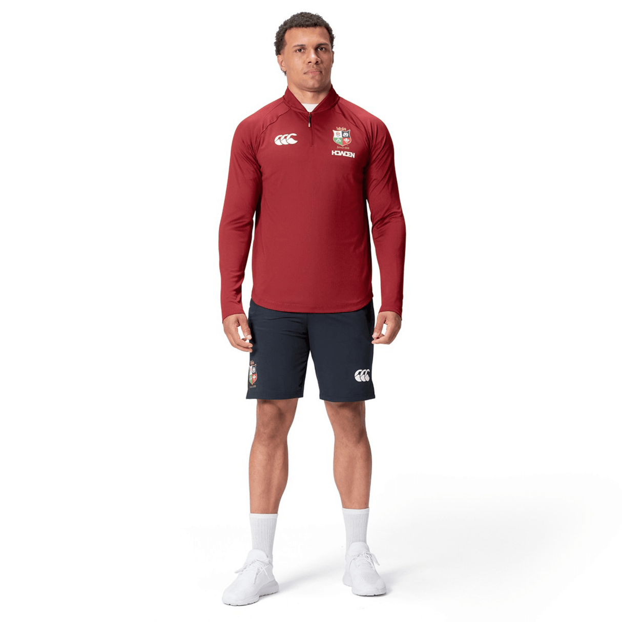 A person wearing a red long-sleeve British & Irish Lions 25 Everest First Layer Top by Canterbury, black shorts, white socks, and shoes stands against a plain white background.