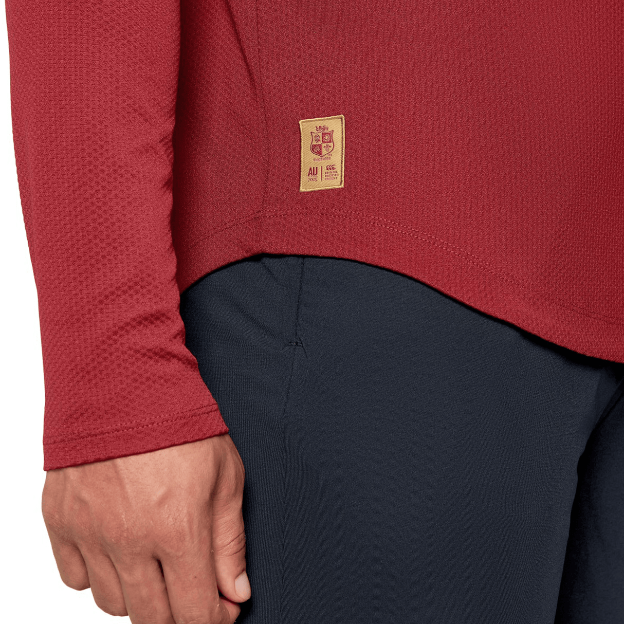 Close-up of a person wearing a British & Irish Lions 25 Everest First Layer Top by Canterbury in red featuring a small patch, paired with black pants. The person's left hand is partially visible.