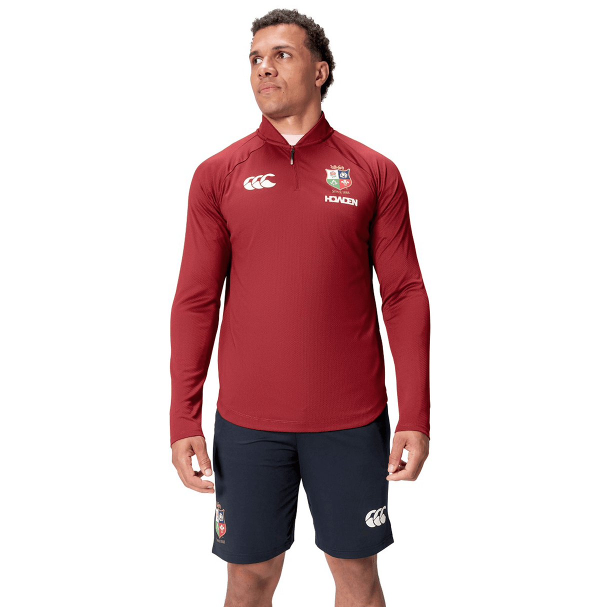 A person wearing a vibrant red British & Irish Lions 25 Everest First Layer Top by Canterbury, adorned with logos, and black shorts stands confidently against a white background.